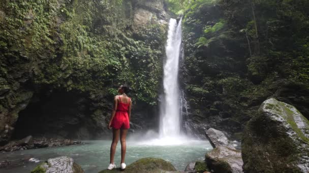 Turista feminino de pé ao pé de uma cachoeira apreciando a paisagem . — Vídeo de Stock