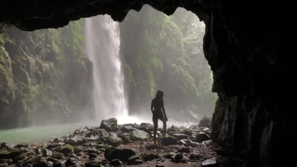 Majestuosa cascada y una cueva oscura con una mujer caminando sobre una superficie rocosa . — Vídeos de Stock