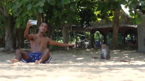 Toeristische man zit op het zand selfie te maken met de aap aan het strand. — Stockvideo