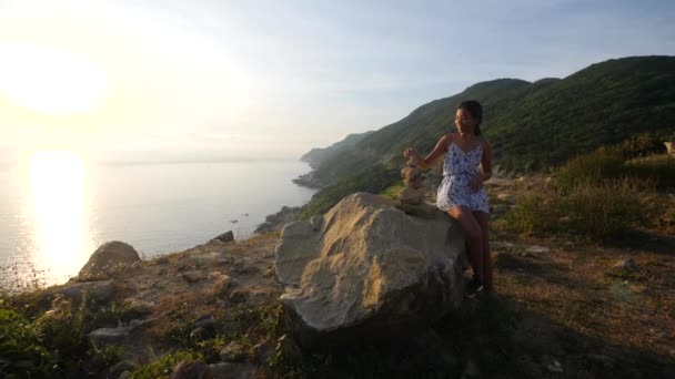 A young woman sitting on a big rock and carefully banlancing stones at sunrise. — Stock Video