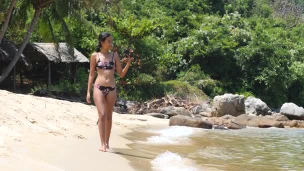 Seorang wanita seksi dengan bikini berjalan dan merekam video dengan smartphone di pantai. — Stok Video