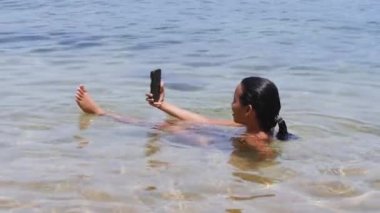 Sahilde sığ sularda oturan bir kadın akıllı telefonuyla selfie çekiyor..