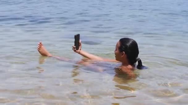 Kvinna sitter i grunt vatten på stranden med selfie video med smartphone. — Stockvideo