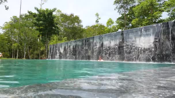 Man och kvinna badar under vattenfallet kaskad i en pool utomhus. — Stockvideo