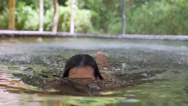 Mladá asijská žena plave ve vodě pod vodní tříšť fontány ve venkovním bazénu. — Stock video