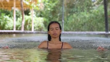 Yüzme havuzunda oturmuş, çeşmenin altında meditasyon yapan genç bir kadın..