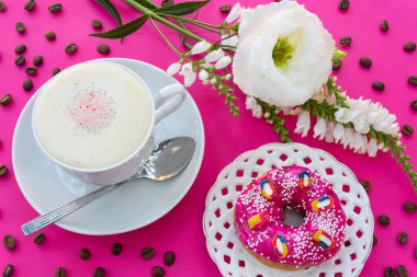 Bir fincan kahve, bir çörek ve kahve çekirdekli parlak pembe bir zemin üzerinde beyaz bir çiçek, parlak kahve molası kavramı, parlak ve hafif bir kahve tadı..