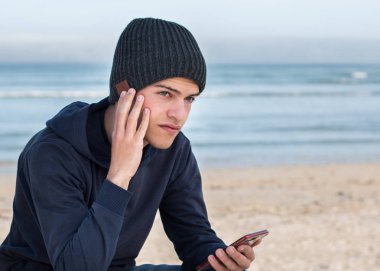 Erkek model içinde Bluetooth hoparlörler ile koyu gri örme bere giyiyor, cep telefonu ile müzik dinlerken açık havada. Düğmelerde parmaklar.