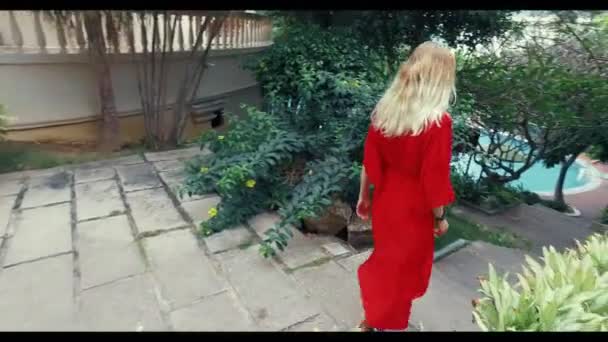 Una joven con el pelo rubio en un vestido rojo baja las escaleras de piedra en el hotel. Baja a la piscina del hotel. A cada lado hay palmeras. La cámara se mueve con un steadicam . — Vídeo de stock