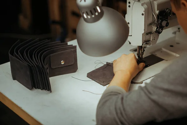 白いテーブルで職人がミシンで革財布を作る。近くにはいくつかの既製の革財布です。テーブルの上にランプとミシン. — ストック写真