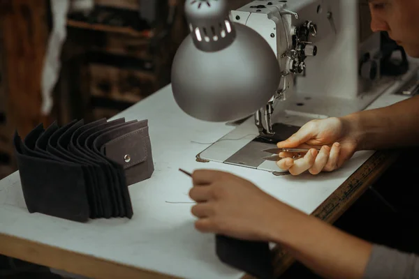 Making leather wallets on a sewing machine by a craftsman on a white table. Nearby are several ready-made leather wallets. On the table is a lamp and a sewing machine.