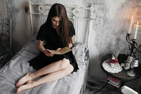 Ένα κορίτσι διαβάζει ένα βιβλίο σε ένα κρεβάτι με ένα ρομαντικό μαύρο φόρεμα. — Φωτογραφία Αρχείου