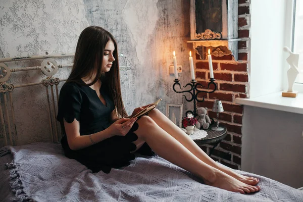 Una ragazza sta leggendo un libro su un letto con un romantico vestito nero . — Foto Stock