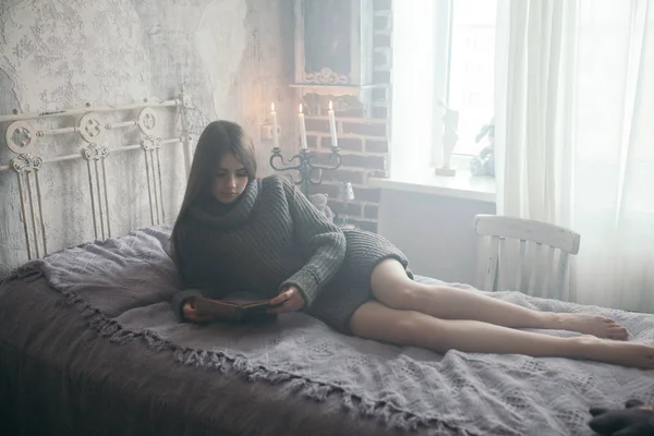 Ung vacker kvinna i varm tröja läsning bok på sängen. — Stockfoto