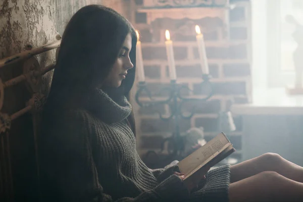 Bella giovane donna in maglione lavorato a maglia seduta sul letto e libro di lettura vicino alla finestra a casa. Atmosfera invernale — Foto Stock