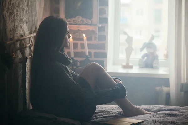 Piękna młoda kobieta z dzianiny sweter siedzi na łóżku i patrząc na okno w pokoju. — Zdjęcie stockowe