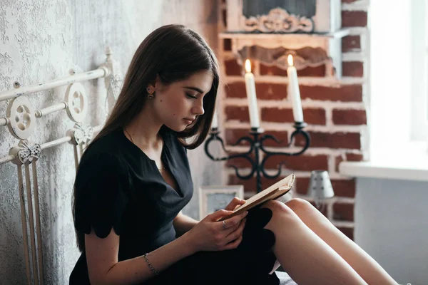 En flicka läser en bok på en säng i en romantisk svart klänning. — Stockfoto