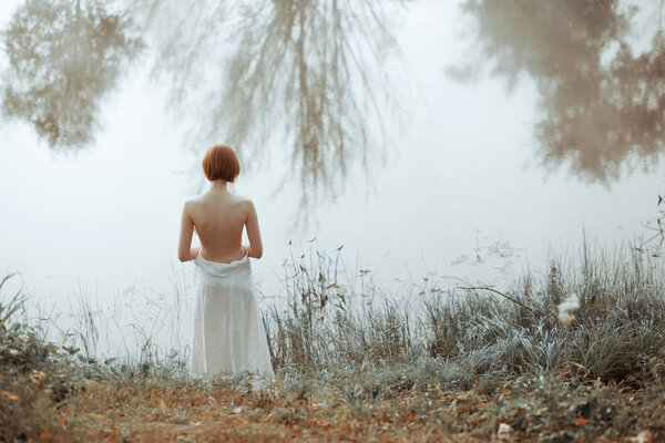 Half-naked attractive woman with short red hair standing in front of specular foggy lake