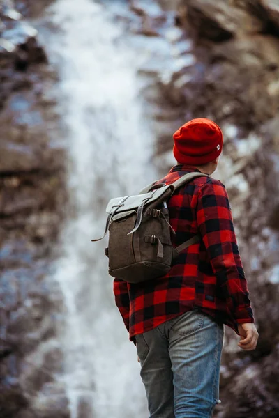 Hipster en gorro rojo mirando la cascada de montaña fría — Foto de Stock