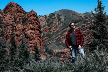 Kırmızı gömlekli genç adam kayaların önünde duruyor