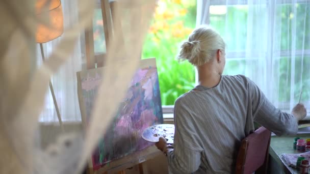 Una ragazza dai capelli biondi sta dipingendo un quadro in una casa di campagna di fronte alla finestra. La finestra mostra un giardino con fiori — Video Stock