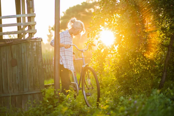 En flicka i en kontrollerad skjorta står med en cykel mot bakgrunden av solnedgången — Stockfoto