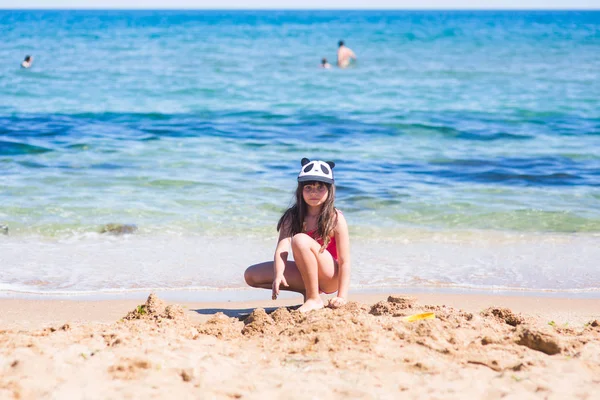 小女孩在一个红色的泳衣和熊猫帽在海滩上建立一个沙堡 — 图库照片