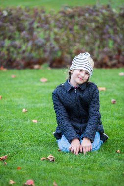 Kız bir siyah ceket ve kot sonbahar yaprakları arasında yeşil bir Çimenlikte oturan