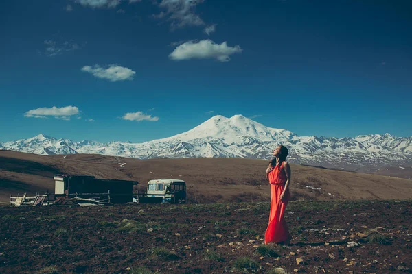 brunette slim girl with closed eyes in long red dress on the background of high snow-capped mountains