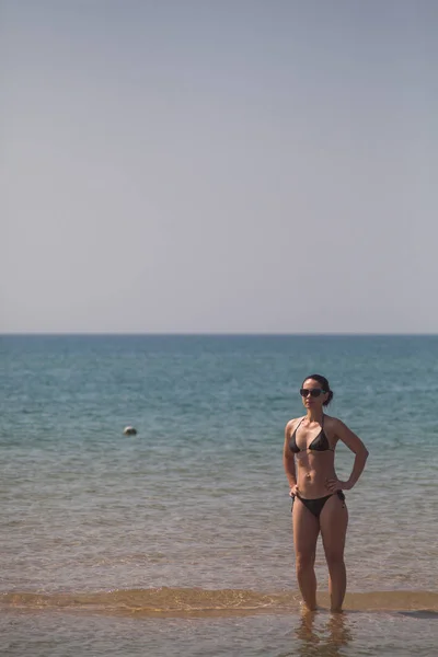 黑发女孩与一个美丽的身影 穿着黑色比基尼和太阳镜站在腰部的腰在海脚踝深在水中的沙滩上的沙滩上 在沙滩上 — 图库照片