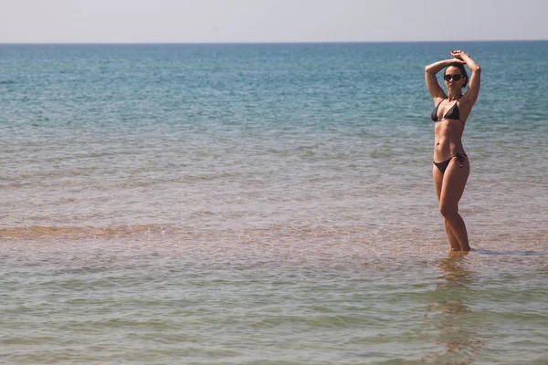 黑发女孩与一个美丽的身影 在黑色比基尼和太阳镜站在半轮在海脚踝深在水中的沙滩上的沙滩上的沙滩上 — 图库照片