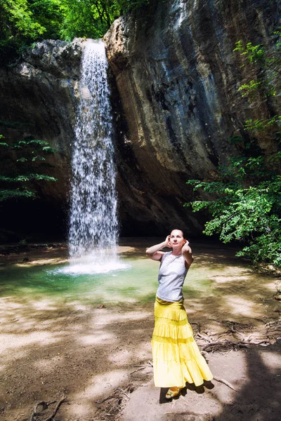 黑发女孩在黄色裙子站在瀑布下的悬崖 — 图库照片