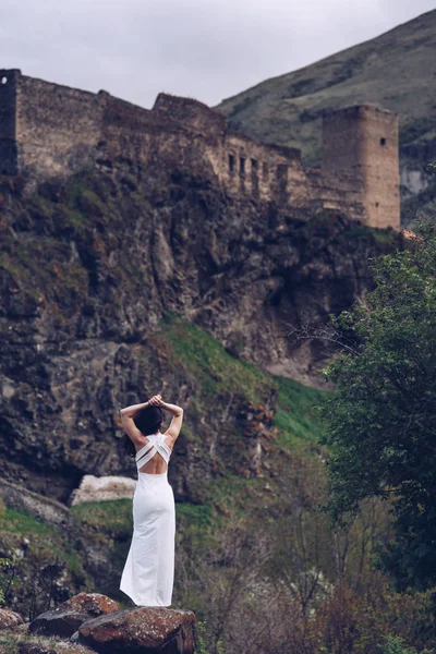 黑发女孩站在她的背部在岩石上一个古老的堡垒的背景白色长裙 — 图库照片