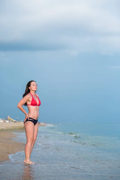 穿着比基尼的纤细的黑发女人站在沙滩上靠近冲浪的地方 她的手放在腰部 — 图库照片