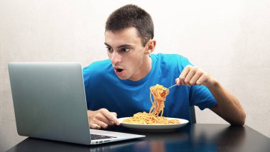 an young man eating spaghetti with tomato sauce and watching the computer clipart