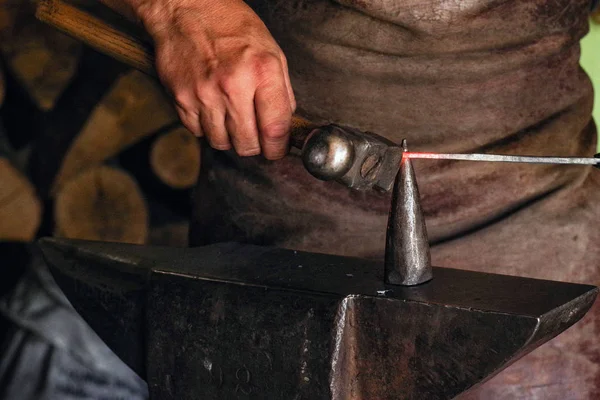 A man in work clothes works with metal. A man holds a hammer and hot metal in his hand. Hot metal .