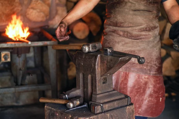 Un hombre con ropa de trabajo trabaja con metal. Un hombre sostiene un martillo y metal caliente en su mano . — Foto de Stock