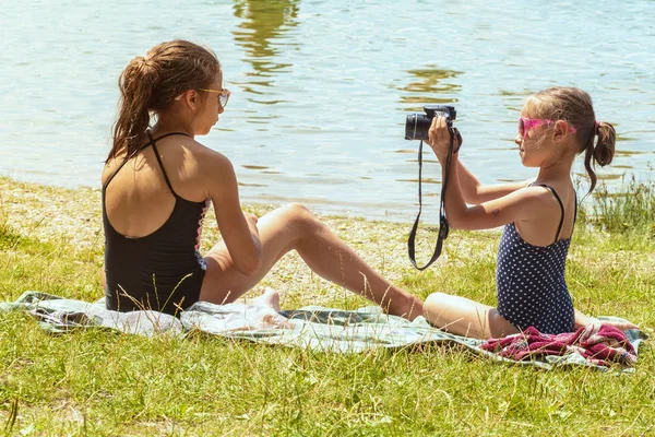 Schattig klein meisje zittend op het gras op zonnige zomerdag en het nemen van foto met camera. Zomervakantie. Klein meisje Foto's nemen zittend in de buurt van de rivier op het gras. — Stockfoto