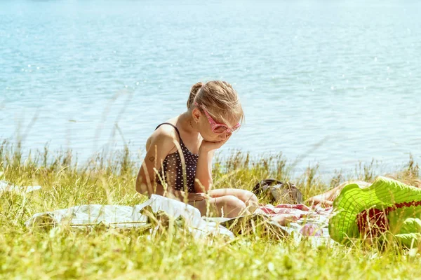 Una chica en traje de baño se sienta sola en la playa en la hierba cerca del río en un día soleado. La chica está triste en la playa. . — Foto de Stock