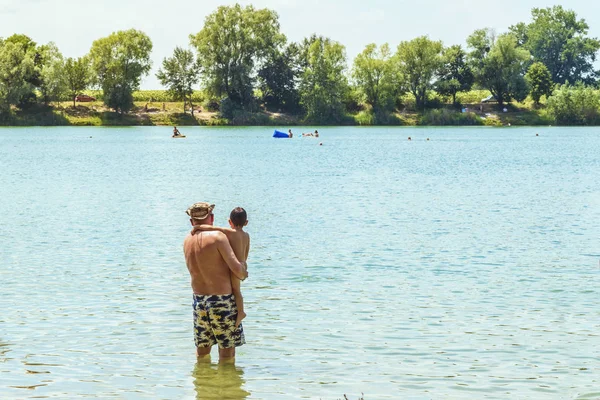Un hombre sostiene a un niño en sus brazos cerca del río. ver el río, familia feliz, padre y su hijo de cinco años cerca del lago al aire libre en un día de verano . — Foto de Stock