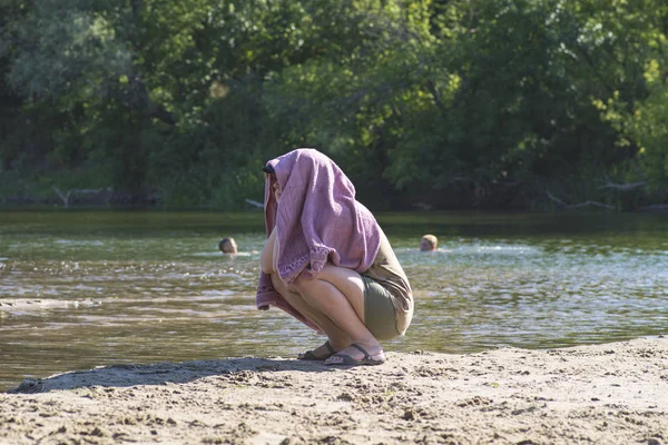 Pigen sidder på stranden med et håndklæde på hovedet. Pigen gemte sit ansigt fra den lyse sol under et håndklæde . - Stock-foto