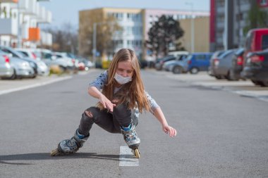 Koronavirüs salgını. Tıbbi maskeli bir kız sokakta paten kayıyor. Tıbbi maskeli küçük bir kız..