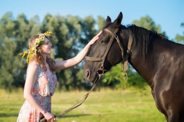 Güzel elbise ve yaz ortası çelenk alan çiçekli giyen genç kadın kahverengi at ile çayır üzerinde yürüme