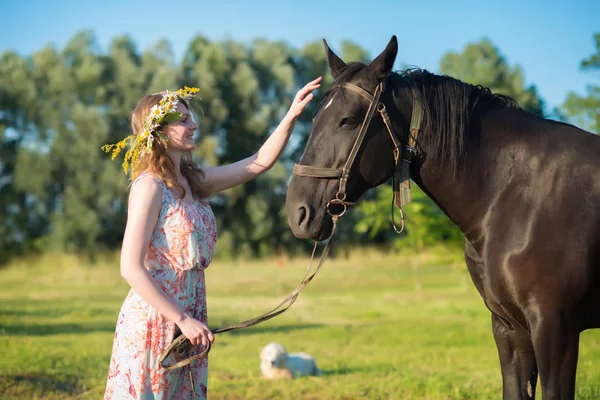 美しいドレスと野の花と真夏の花輪を身に着けている牧草地に茶色の馬で歩く若い女性 — ストック写真