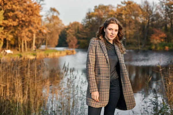 Serious long-haired woman wearing brown coat and standing at autumn park at a lake, windy — Stock Photo, Image