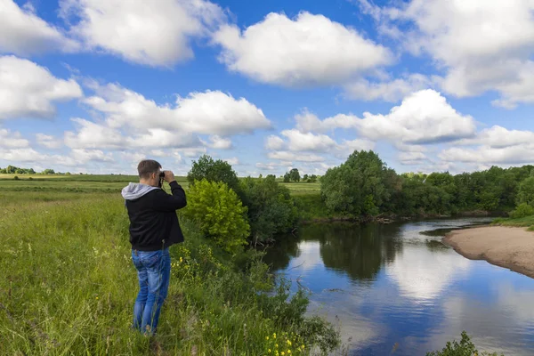 A man stands on the bank of a river and look through binoculars.