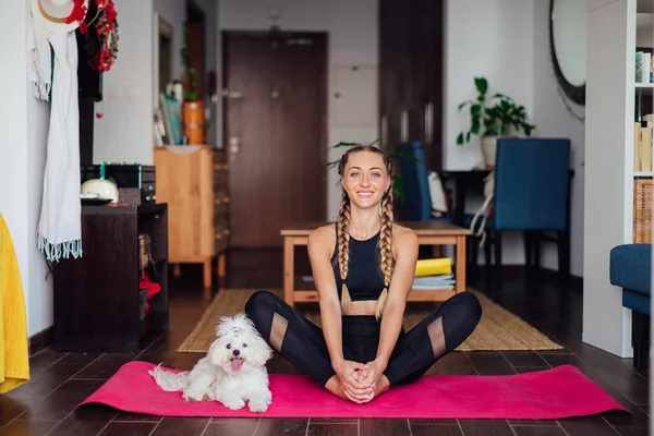 Mulher bonita fazendo exercício, praticando ioga, cachorrinho branco bonito senta-se ao lado de seu proprietário, menina esportiva vestindo sportswear preto trabalhando fora dentro de casa Imagem De Stock