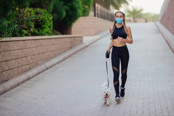 Mulher desportiva usa uma máscara facial correndo junto com seu cão dentro das ruas da cidade . Fotografias De Stock Royalty-Free