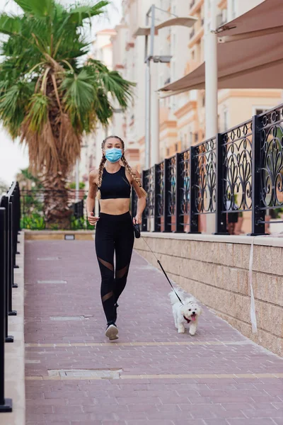 Mulher desportiva usa uma máscara facial correndo junto com seu cão dentro das ruas da cidade . Fotografia De Stock