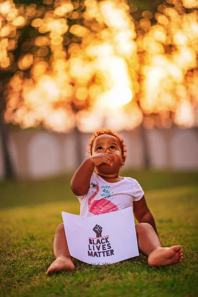 Pequena menina afro-americana senta-se na grama verde e segura o cartaz Black Lives Matter Fotografias De Stock Royalty-Free